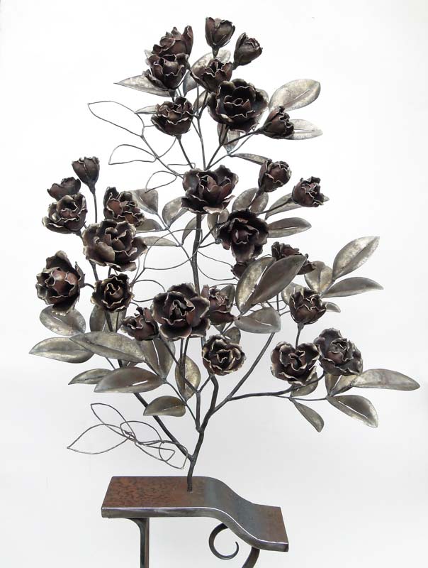 バラの立木をモチーフにした鉄の鍛造作品です。一枚一枚の花びらを鍛造してガス溶接で組み上げています。 花は「シャルル・アズナブール（マチルダ）」をモデルに、一部の葉はワイヤー表現。 軽やかで、風で動くような佇まいを意図したアイアンアート。台は少し装飾的なデザインになっています。