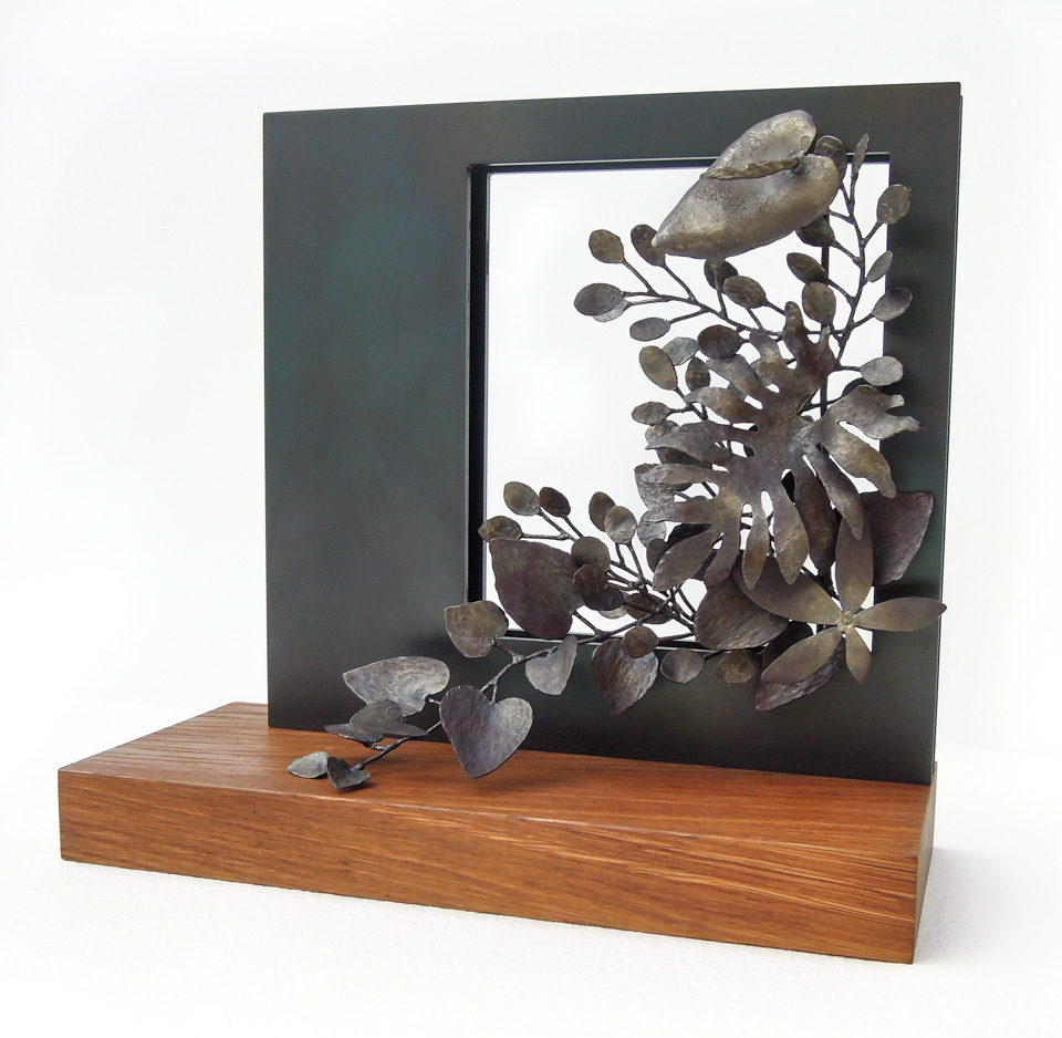 四角い窓から様々な植物が溢れでているようなアートオブジェです。窓と植物は鉄、ベースは木でできています。斜めからの写真。