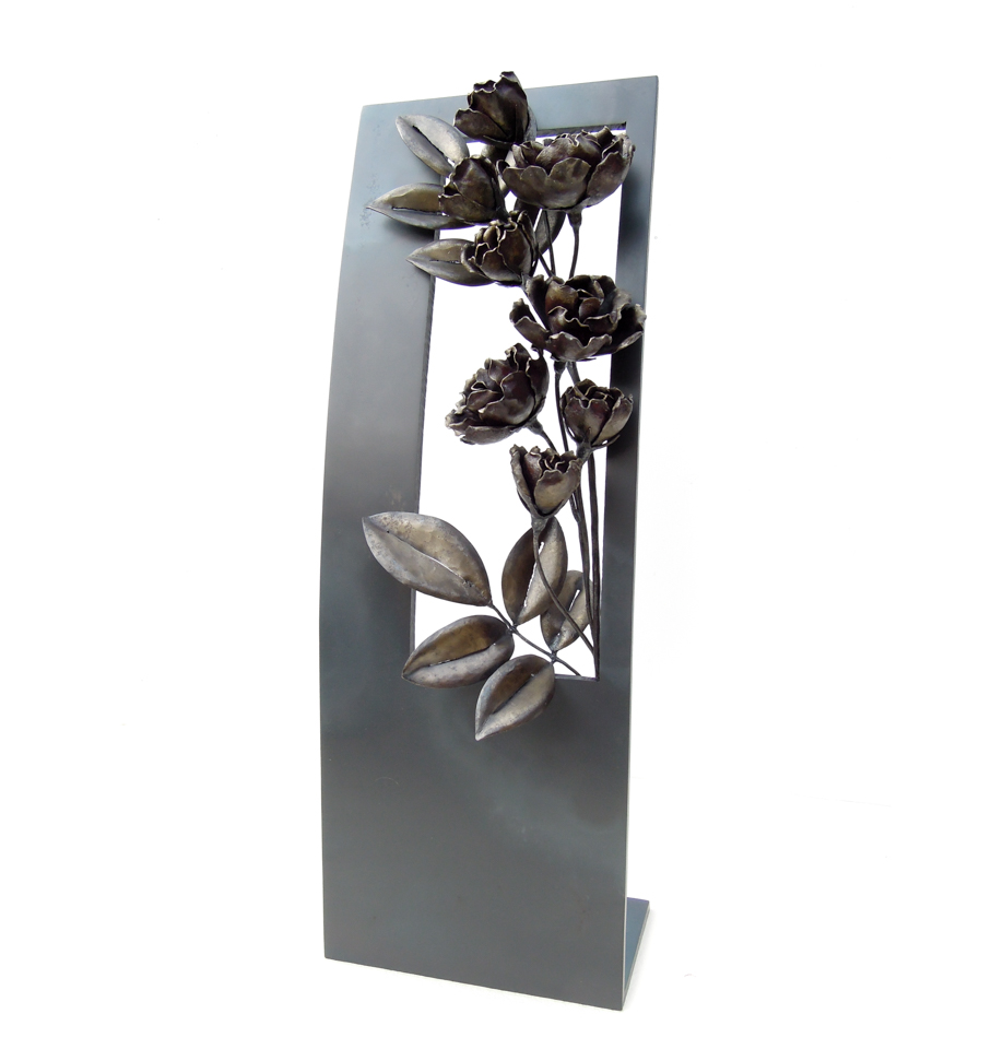 縦長の窓から一枝のバラが咲き誇っているアートオブジェです。窓はゆるやかな曲面になっており、花付きの良い枝がいきいきと伸びています。フレーム、バラともに鉄で制作し、同じ素材で色味の違いを出しています。斜めからの写真。