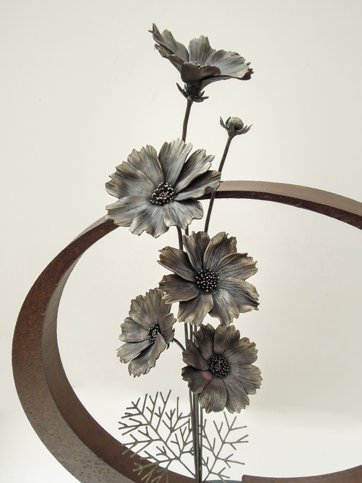 円の鉄プレートに鍛鉄のコスモスの花が添えられた彫刻作品です。コスモスの花の表現。蕾から咲き誇る状態まで変化のある表現です。花の中心部に熱による色の変化が見えています。